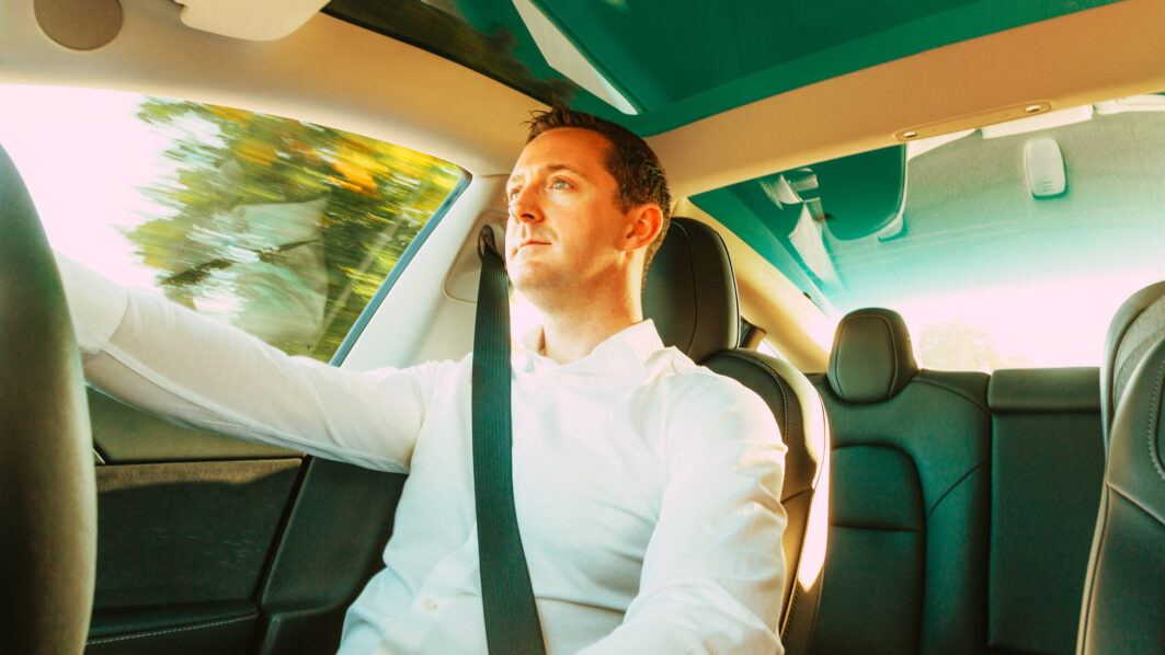 Wide angle color image of a mid adult man in his 30s driving inside a modern electric car. He wears a smart white shirt on his way home through the city. Motion blur on the scenery as the car speeds past.